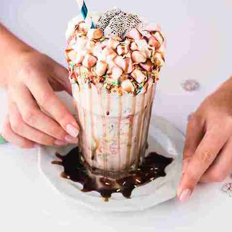 A rich chocolate milkshake overflowing with whipped cream, mini marshmallows, sprinkles, and chocolate drizzle, served in a tall glass with a paper straw and hands holding the plate.