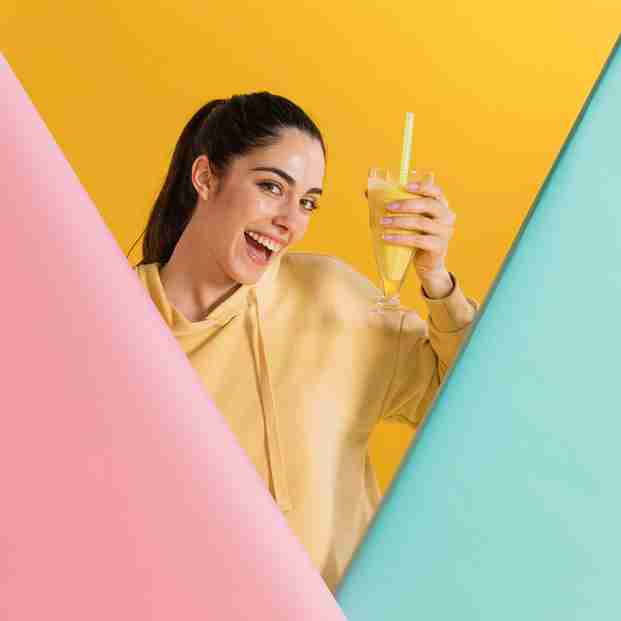 A smiling woman in a yellow hoodie holding a glass of yellow smoothie with a straw, standing against a vibrant background of yellow, pink, and turquoise colors.
