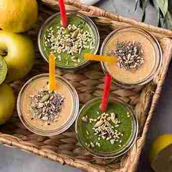 Four colorful smoothies—two green and two beige—served in clear glasses with red and yellow straws, topped with nuts and seeds, placed in a woven basket alongside fresh apples and lemons.
