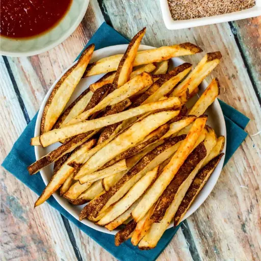 Top-down view of crispy skin-on French fries served on a white plate with a side of ketchup and seasoning salt on a rustic wooden table.